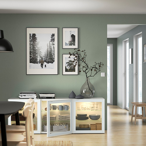 Modern dining area with BESTÅ sideboard featuring glass doors, showcasing dishes and decorated with framed artwork and a glass vase with branches.