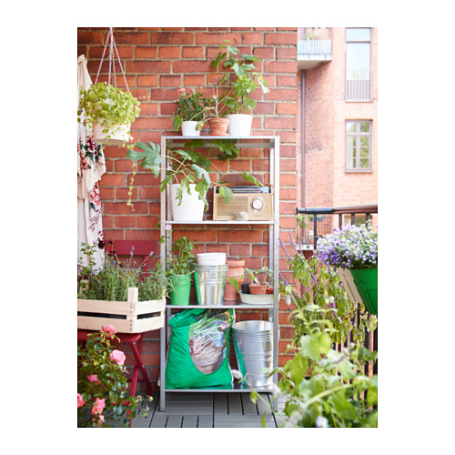 Estantería de balcón con plantas en macetas, contra pared de ladrillo rojo. Estantes de metal, macetas blancas y verdes, radio vintage y mesa de exterior.