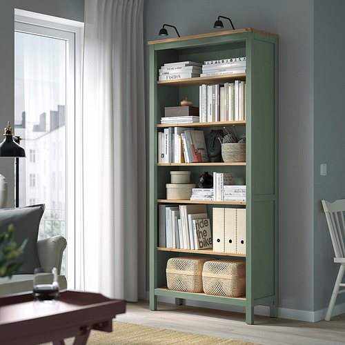 Librería HEMNES en color verde grisáceo con estantes de madera, que sostienen libros y decoración en una habitación acogedora.