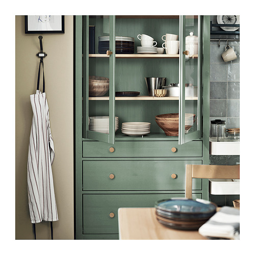 Mueble de madera HEMNES verde con puertas de cristal, donde se exhiben platos y cuencos. El delantal cuelga en el lado izquierdo, la encimera de la cocina en primer plano.