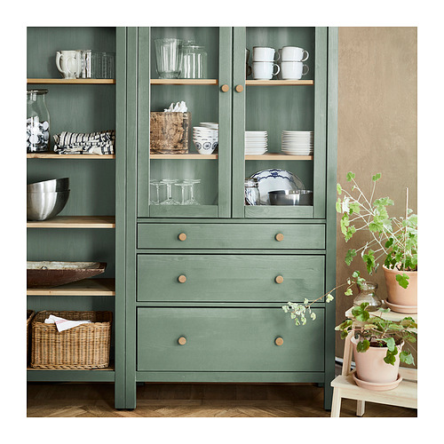 Un armario de madera verde con puertas y cajones de cristal. Sus estanterías están repletas de platos y vasos, con cestas de mimbre y plantas que decoran el espacio.