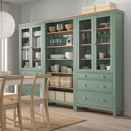 Mueble de almacenaje HEMNES verde, puertas de cristal, estanterías de madera, cuencos, vajilla, copas de vino, libros, sillas.
