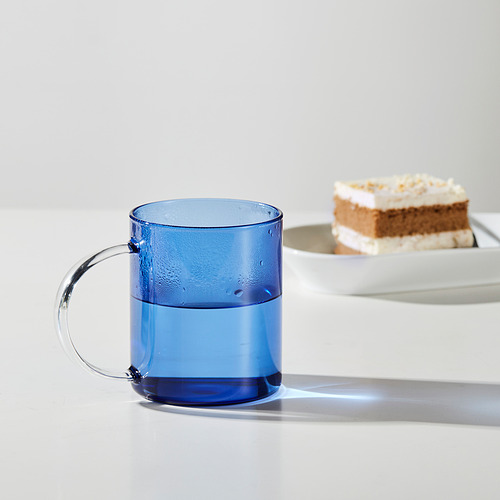 Taza de cristal azul con asa y medio llena de agua, está junto a una porción de pastel en una bandeja blanca.
