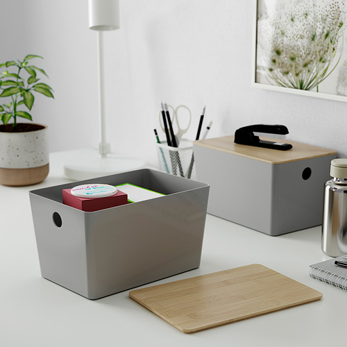 Pequeñas cajas KUGGIS grises sobre un escritorio blanco. Uno está abierto, revelando elementos coloridos en el interior. La caja cerrada encaja perfectamente debajo de una lámpara y al lado de una planta.
