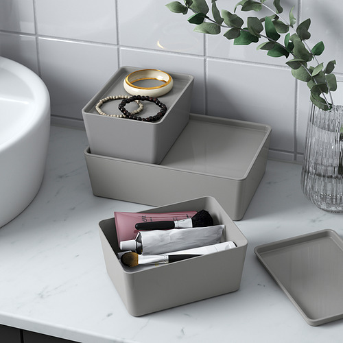 Tres cajas KUGGIS grises sobre la encimera de un baño. Uno contiene maquillaje, otro joyas y otro está vacío. También hay un jarrón con vegetación.