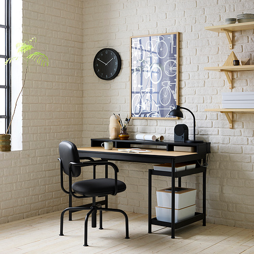 Moderna configuración de escritorio MULLSJÖ negro contra pared de ladrillo blanco, que incluye silla negra, escritorio de madera con almacenamiento y reloj de pared único.