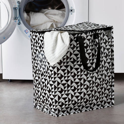 La cesta de ropa geométrica en blanco y negro de KNALLA contiene muchos artículos, colocados junto a una lavadora blanca más grande.