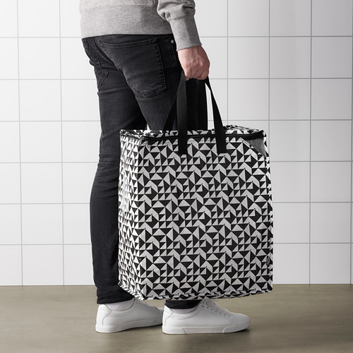 Persona sosteniendo una gran bolsa de compras KNALLA con patrón geométrico. La bolsa tiene asas y parece resistente para hacer la compra o lavar la ropa.