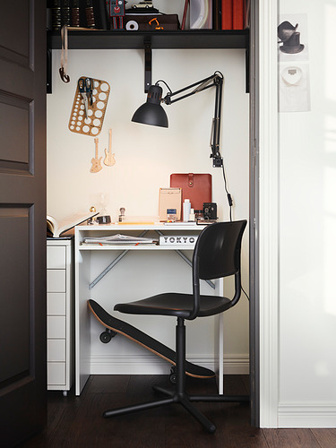 Pequeño escritorio negro, TORALD, diseño minimalista, superficie de melamina, cajón para almacenaje, silla negra, lámpara de pared, espacio de trabajo organizado.