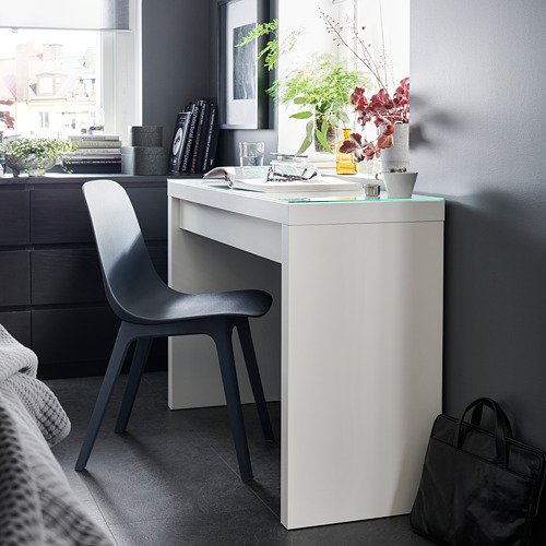 Moderna silla negra junto a un escritorio blanco con tapa de cristal, con un espacio de trabajo cuidadosamente dispuesto con decoración y libros en un elegante ambiente.