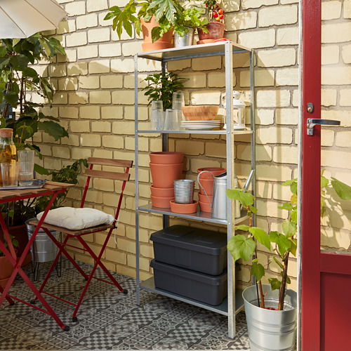 Configuración de patio con estantes de plata HYLLIS que sostienen plantas, contenedores y platos. Sillas rojas y fondo de pared de ladrillo.