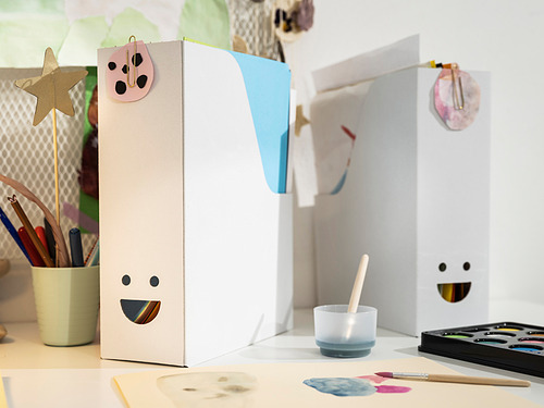 Dos archivos blancos y sonrientes de la revista TJABBA hechos de cartón corrugado 100% reciclado sobre una mesa con materiales de arte.
