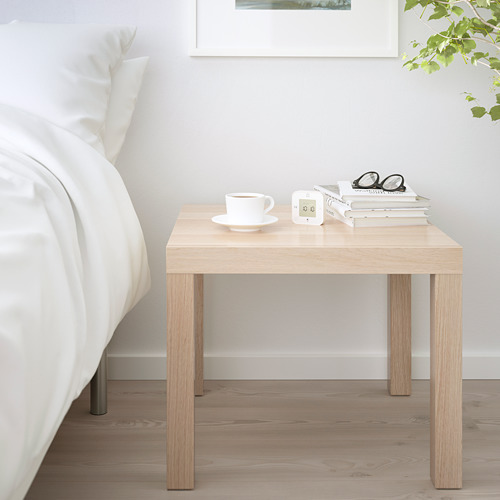 Mesa auxiliar LACK como mesita de noche al lado de la cama, sosteniendo taza, libros, vasos, complementando la ropa de cama blanca en el dormitorio minimalista.