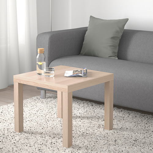 Mesa auxiliar de roble LACK junto a sofá gris con vidrio, jarra y revistas sobre alfombra blanca.