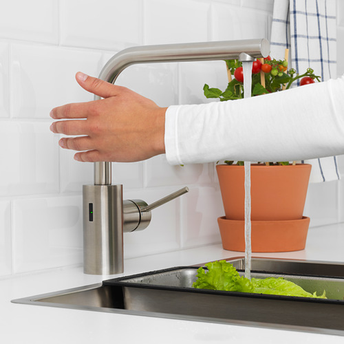Persona lavándose las manos bajo el grifo de acero inoxidable con agua fluyendo hacia el fregadero de la cocina; Plantas en macetas cercanas.