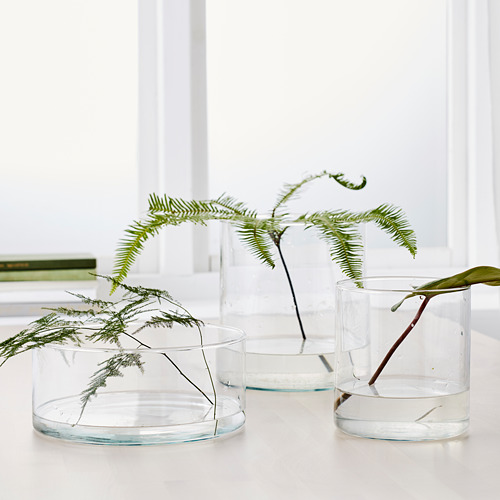 Tres recipientes de vidrio de varios tamaños, con plantas verdes, dispuestos sobre una mesa blanca. Sirve como jarrones y organizadores.