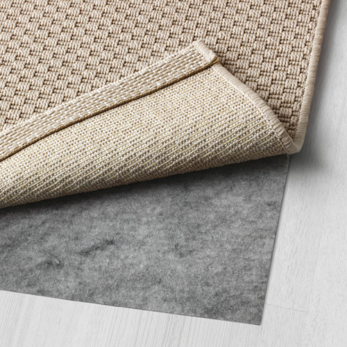 Alfombra tejida de color beige claro plegada sobre una capa inferior antideslizante de color gris oscuro, expuesta sobre un suelo de madera clara.