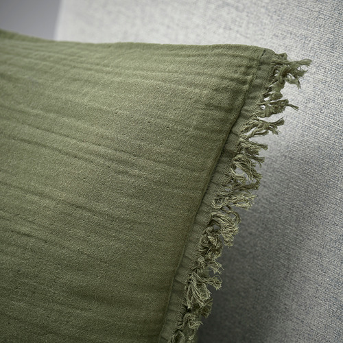 Primer plano de la funda de cojín VALLKRASSING verde y suave con bordes limpios y deshilachados sobre textil gris.