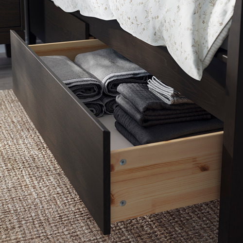 Cajón de madera abierto debajo de la cama que muestra mantas grises cuidadosamente dobladas. La cama tiene un marco de madera oscura, una almohada blanca con estampado floral y una alfombra beige cerca.