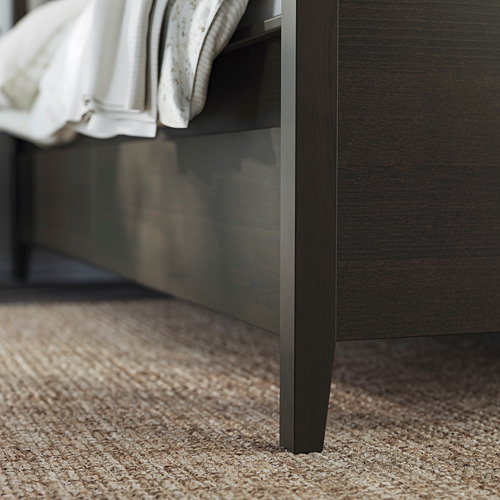 Cama de madera oscura de IDANÄS con sábanas blancas y alfombra beige, con patas elegantes y oscuras y espacio de almacenamiento.