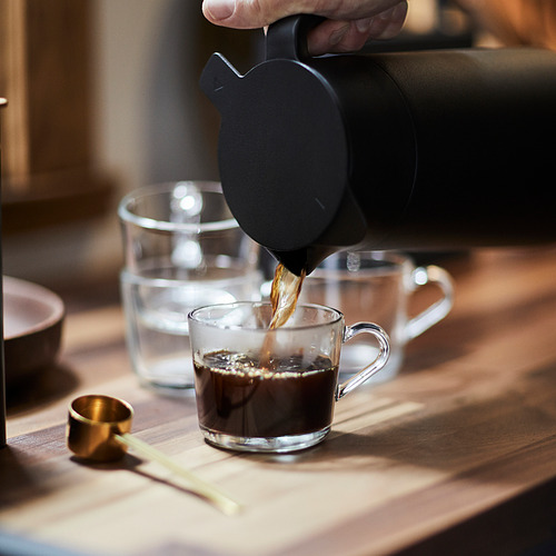 La persona vierte el café en una taza de vidrio transparente sobre una mesa de madera. Taza que forma parte de la serie IKEA 365+, diseñada para ofrecer durabilidad y versatilidad.