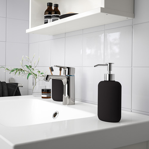 Lavabo de baño moderno con elegante grifo cromado y dispensador de jabón negro, planta pequeña, soporte para cepillos de dientes y frascos en el mostrador, azulejos blancos.