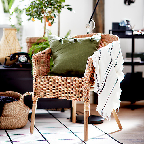 Acogedora escena interior con silla AGEN de mimbre, almohada verde, manta blanca, cestas y plantas.