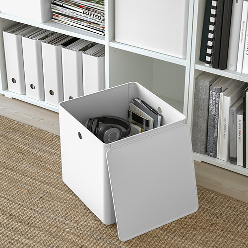 Caja de almacenamiento blanca con tapa, auriculares, papeles, sobre alfombra. Estanterías con libros, carpetas. Serie KUGGIS.