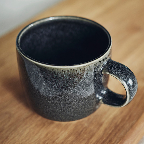 Taza vidriada de color gris oscuro sobre superficie de madera; tacto suave y artesanal, parte de la serie GLADELIG.