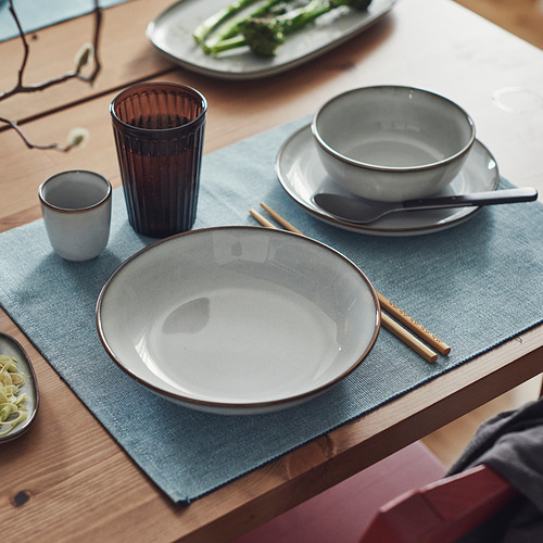 La placa profunda GLADELIG, de color arena y gres, se asienta sobre una estera azul, con palillos, taza y cuchara cerca. Más grande que los elementos circundantes.