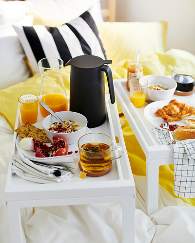Bandeja de desayuno con almohadas blancas; jugo de naranja, medialunas, cereales y un termo de café negro.