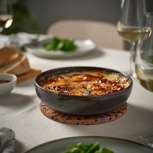 Un pequeño cuenco de gres gris oscuro se asienta sobre un posavasos de corcho, lleno de platos en capas. Rodeado de platos y copa de vino, sobre un mantel ligero.