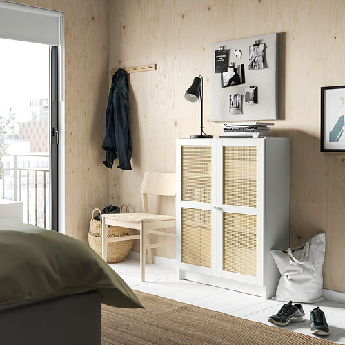 Habitación minimalista con elementos naturales: librería blanca BILLY con puertas de bambú, ropa de cama verde, abrigo negro, escritorio con lámpara y silla de mimbre.