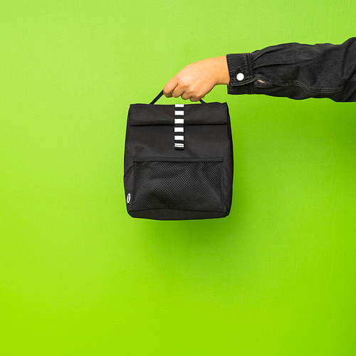 Persona sosteniendo una bolsa de almuerzo rectangular negra con bolsillo de malla contra la pared verde.