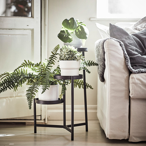 Moderno soporte para plantas de metal negro con tres macetas blancas que sostiene varias plantas frondosas, colocadas junto a un sofá crema con mantas. Decoración interior del hogar.