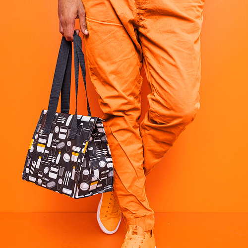 La persona sostiene una bolsa de almuerzo negra estampada sobre fondo naranja. Bolso diseños lúdicos de tazas y botellas.