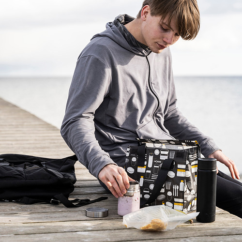 Un hombre se sienta en el muelle, sosteniendo una bolsa de almuerzo gris FLADDRIG con un patrón de temática de café. A su lado, se colocan una mochila negra y una botella.