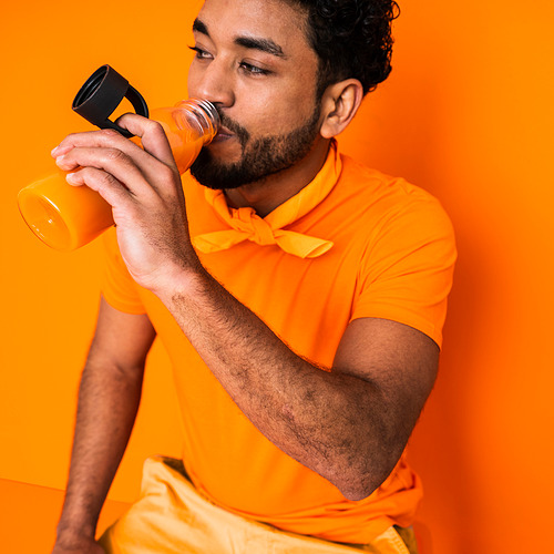 Hombre con camisa naranja bebiendo de una botella transparente con tapa y correa negras, sobre un fondo naranja.