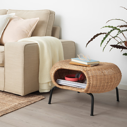 Reposapiés de ratán GAMLEHULT beige con libros y caja roja, junto a sofá con almohadas y planta. Compacto y funcional.