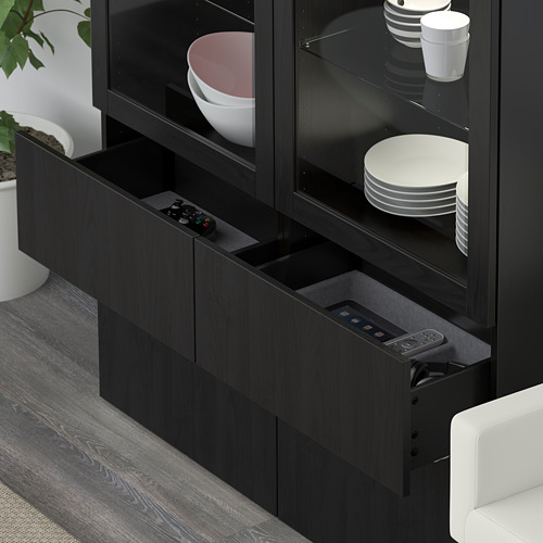 Mueble BESTÅ negro con cajón abierto con mandos a distancia, junto a sofá blanco.