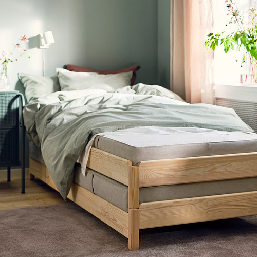 Cama deshecha con estructura de madera clara, sábanas verdes, almohadas blancas, contra la pared verde, plantas cerca.