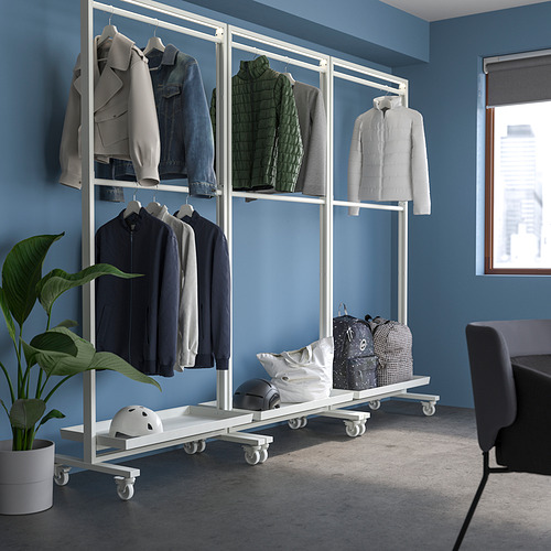 Expositor de perchero rodante MITTZON con ruedas, de color blanco acero, que sostiene varias chaquetas, bolsos, cascos y plantas junto a una silla y ventana grises.