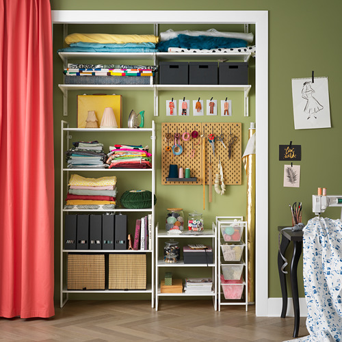 Sala organizada con estanterías de acero blanco JONAXEL, tela verde, ropa doblada, tablero de clavijas, arte y contenedores de almacenamiento.