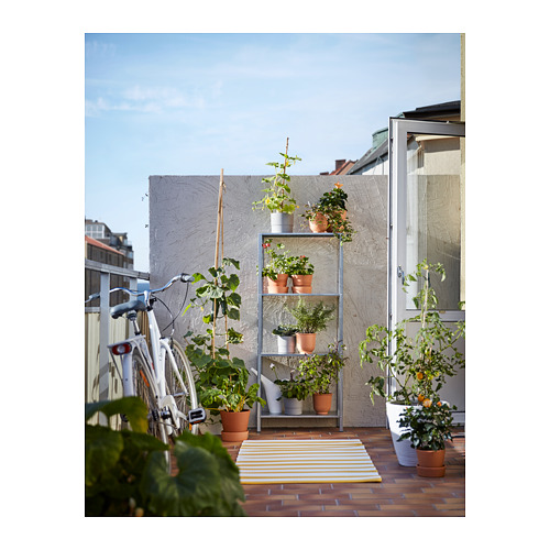 Pequeño balcón con plantas en estanterías HYLLIS, bicicleta blanca, alfombra amarilla, edificios urbanos, cielo despejado.