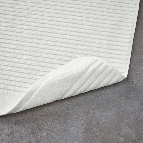 Alfombra de baño blanca con superficie texturizada, dispuesta sobre suelo gris oscuro. Diseñado para un secado rápido y una alta absorción.
