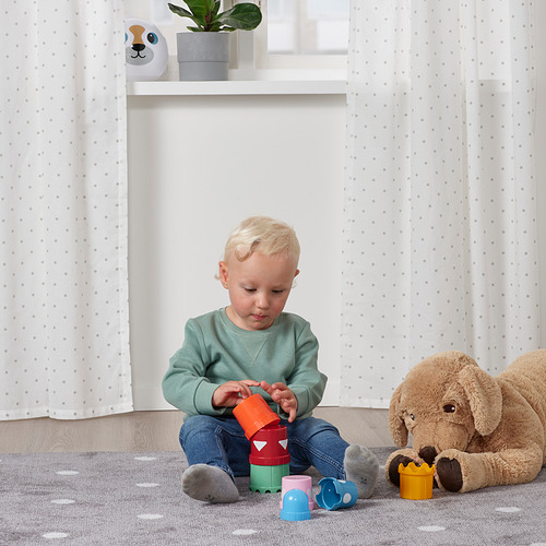 Niño pequeño jugando con coloridas tazas apilables en el suelo, rodeado de peluches.