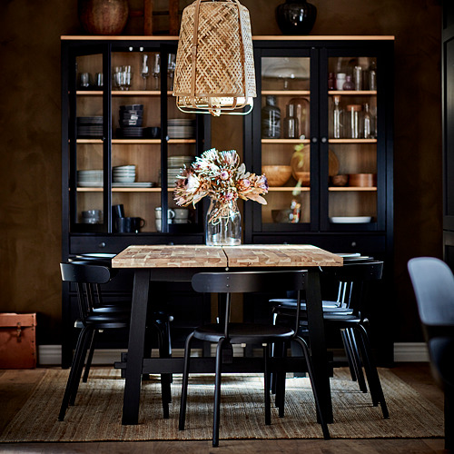 Acogedora configuración de comedor con sillas negras YNGVAR alrededor de una mesa rústica, debajo de un gabinete negro con puertas de vidrio.