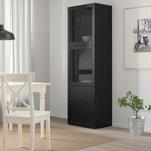 Elegante mueble de almacenaje BESTÅ negro, alto con puertas y cajones de cristal, que alberga utensilios de cocina junto a una silla blanca y una planta.