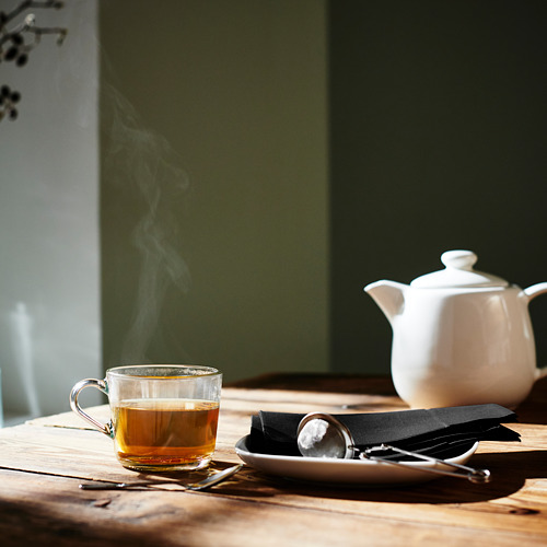 Taza de vidrio con té dorado, vapor ascendente, infusor de té IDEALISK en servilleta junto a tetera blanca sobre mesa de madera.