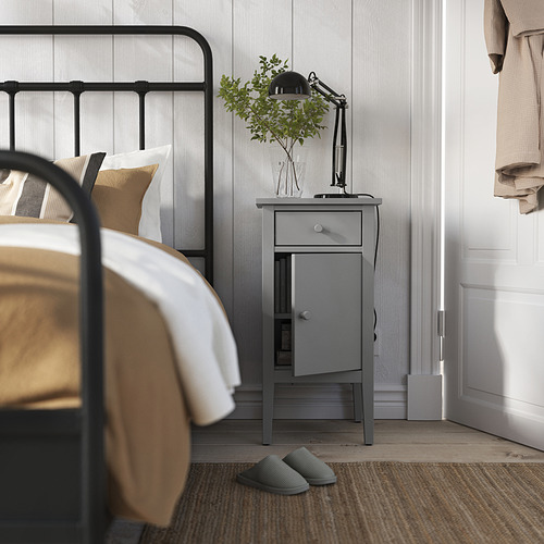 Escena de dormitorio con cama de metal negro, mesita de noche GULLABERG gris, lámpara, planta y zapatillas.